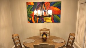 A dining room with a round wooden table, four chairs, a colorful abstract painting above the table, and a chandelier.