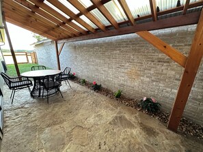 Eine überdachte Terrasse mit Backsteinmauer, Holztisch und -stühlen sowie einem kleinen Blumenbeet mit Pflanzen.