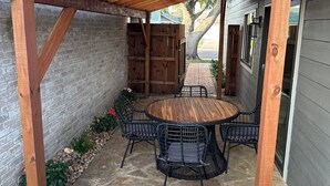 A covered patio with a stone floor, a wooden pergola, a round table with chairs, and a brick wall.