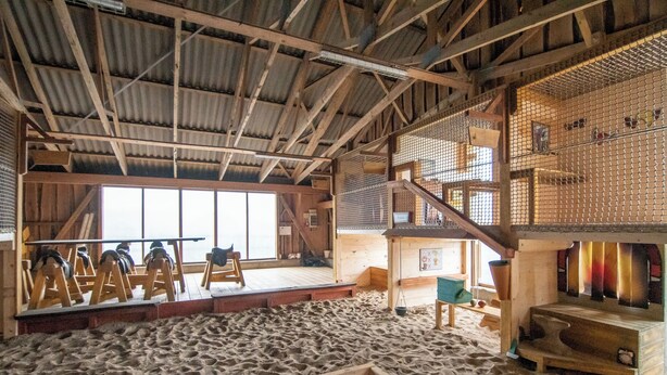 Un espace de jeux intérieur avec un sol sablonneux, des structures en bois et un plafond haut.