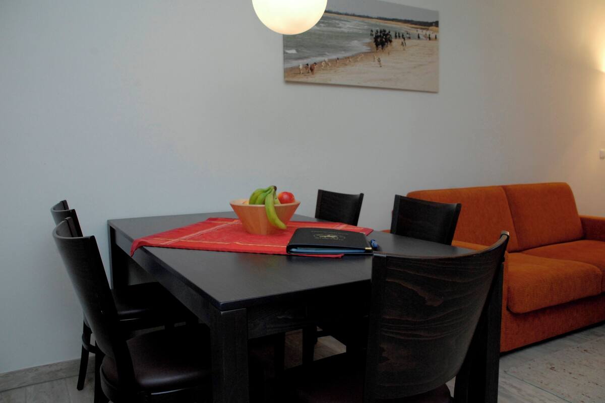 Uma área de refeições com uma mesa, cadeiras, uma tigela de frutas e um jogo americano vermelho.