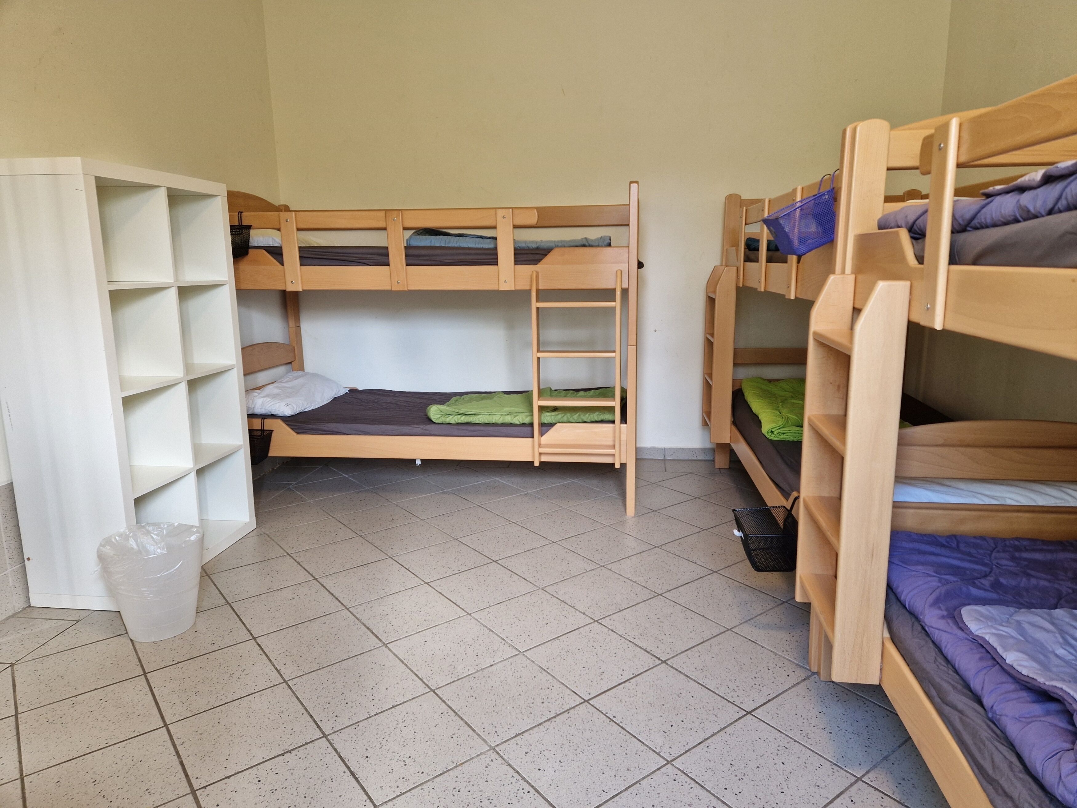 A dormitory room with bunk beds, a white shelving unit, and a tiled floor.