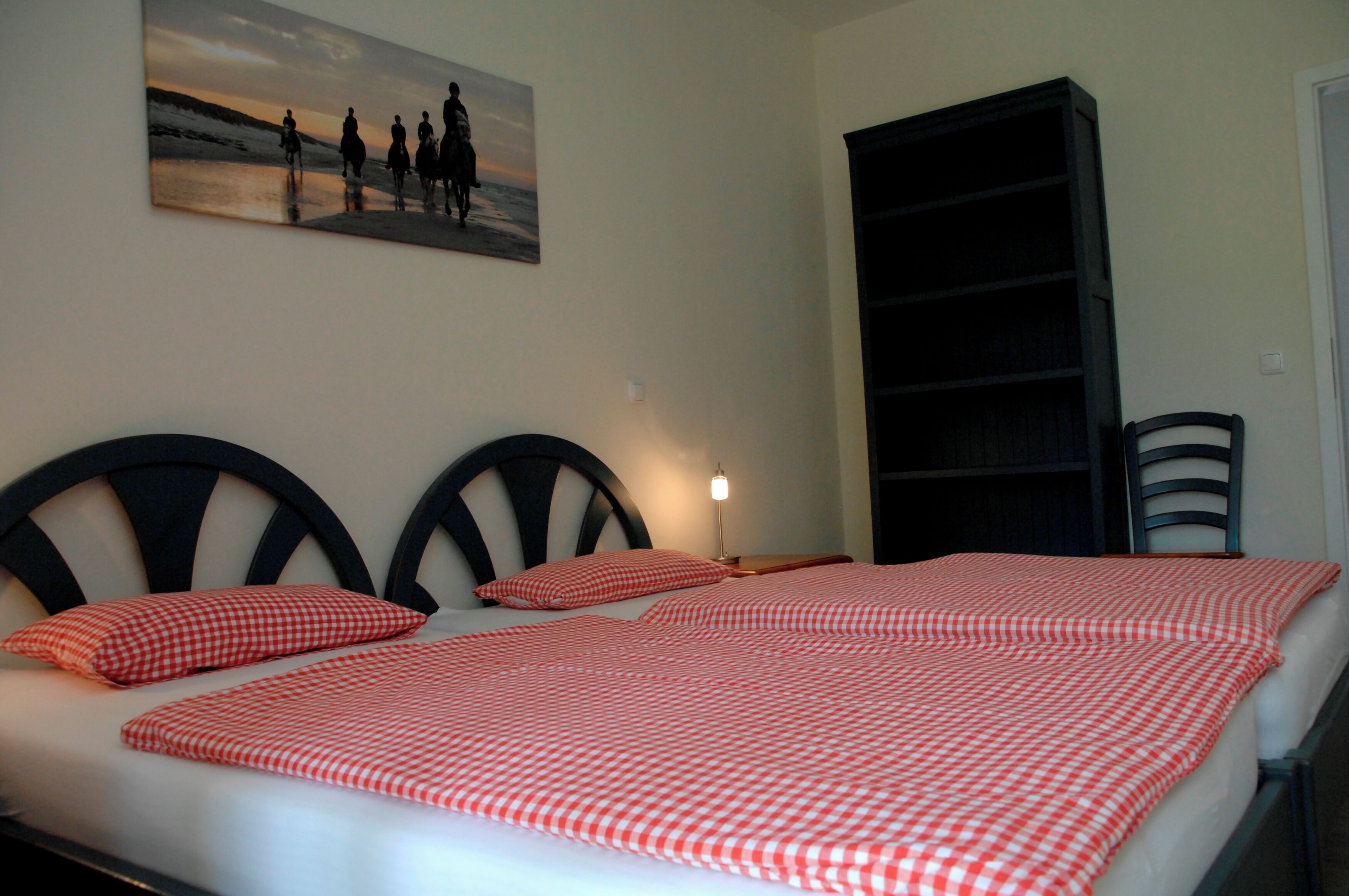 A bed with a checkered bedspread, a painting of people on horseback, a bookshelf, and a chair.