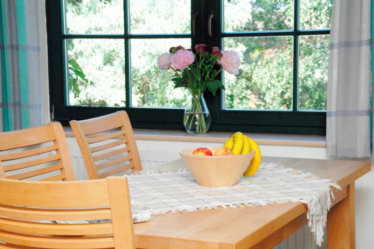 Uma mesa de madeira com uma tigela de frutas e um vaso de flores perto de uma janela.