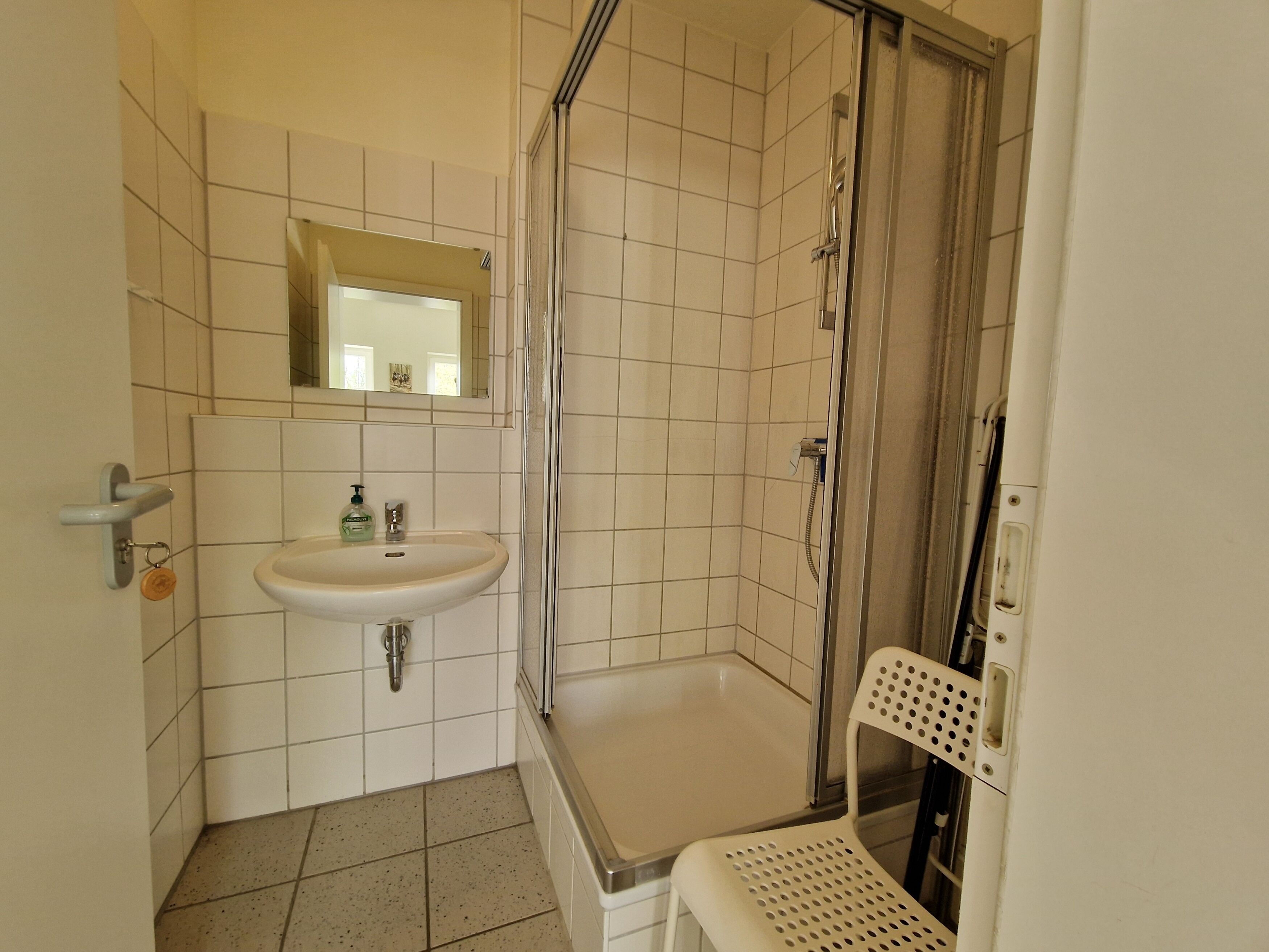 A bathroom with a sink, mirror, shower, and a chair.