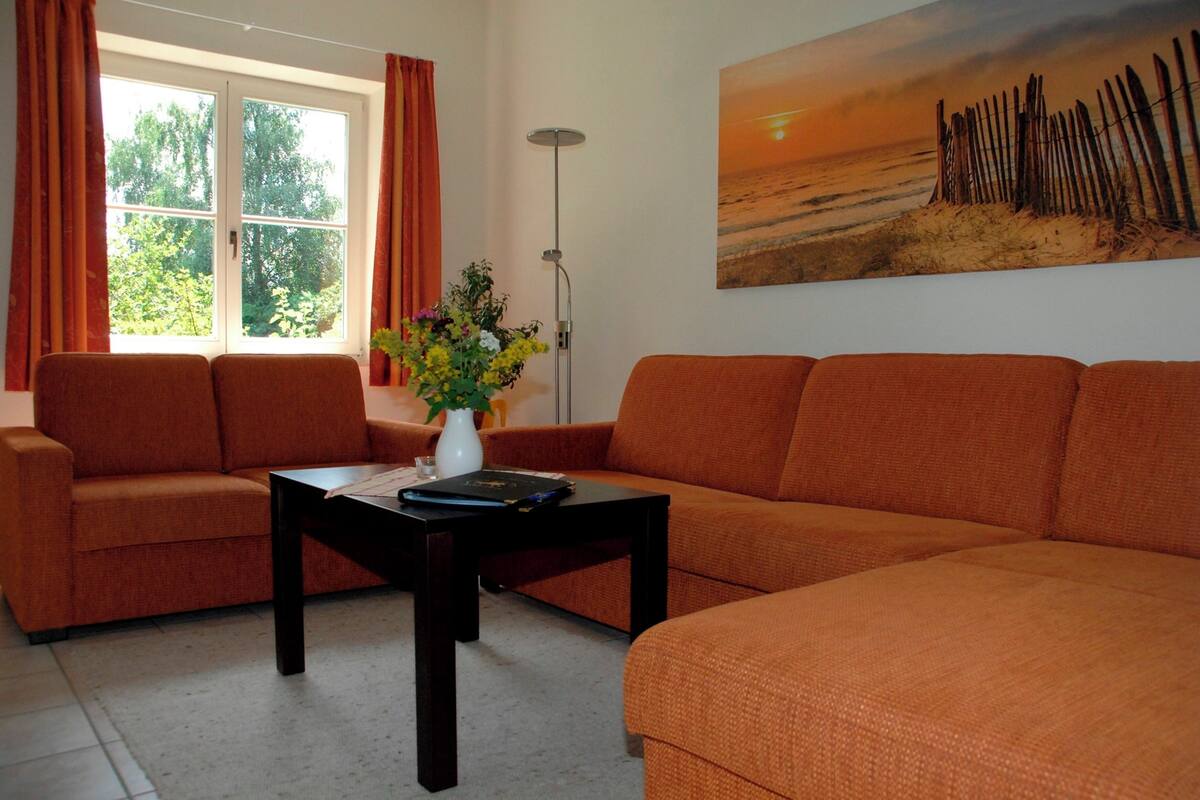 Uma sala de estar com sofás laranja, uma mesa de centro, um vaso com flores e uma pintura de uma cena de praia na parede.
