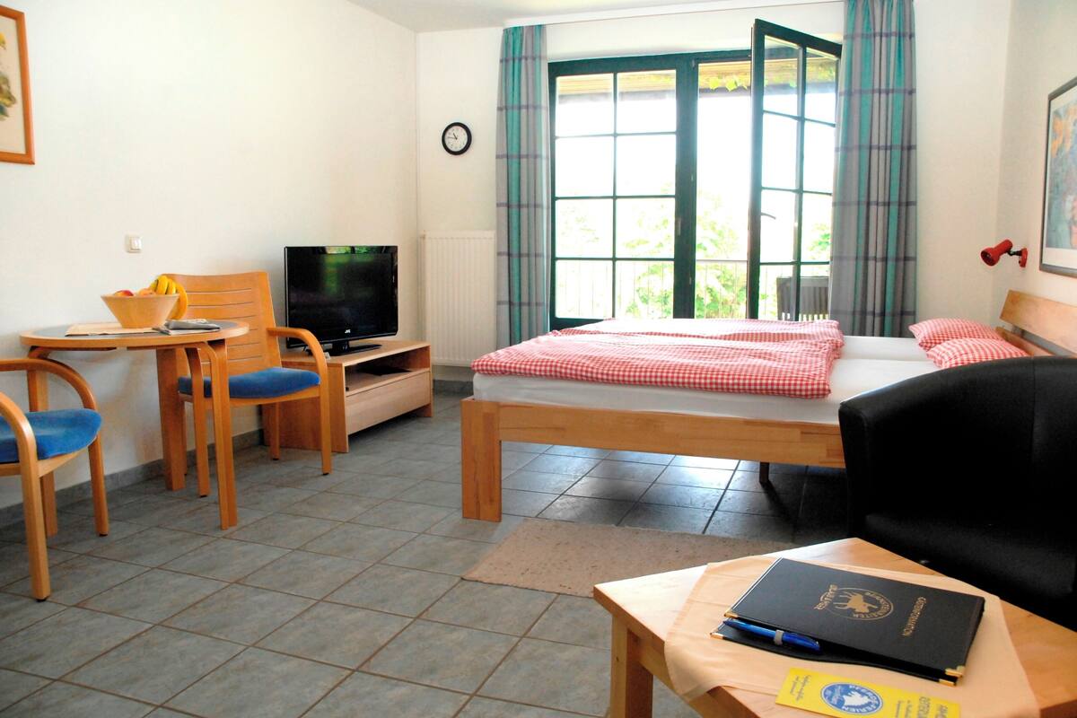 Quarto de hotel com cama, escrivaninha com cadeira, televisão e janela com cortinas.