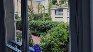 Uitzicht vanuit een raam met balkon, op een tuin met diverse planten en een gebouw op de achtergrond.