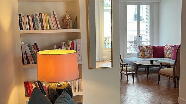 Een woonkamer met een bank, een boekenkast, een lamp en uitzicht op buiten.
