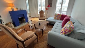 Een woonkamer met een blauwe open haard, houten meubels, een grijze bank met kleurrijke kussens en grote ramen met uitzicht op gebouwen.