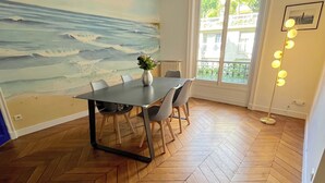 Een eetgedeelte met een visgraatvloer, een tafel met stoelen, een vaas met bloemen, een groot raam met uitzicht op groen en een muur met een zeemuurdecoratie.