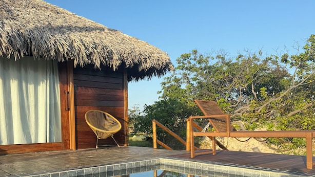 A pool with tiled edges, a wooden deck, a thatched-roof building, and a wicker chair. - Pacas Suítes (Santo Amaro do Maranhão)