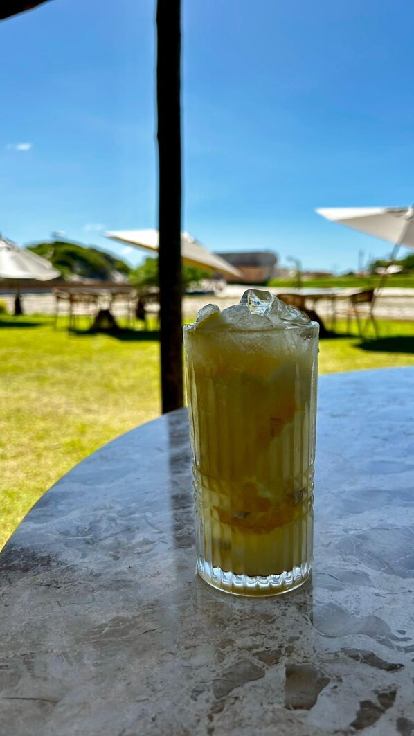 Un vaso de bebida helada sobre una mesa, con vista a un área verde y sombrillas al fondo.