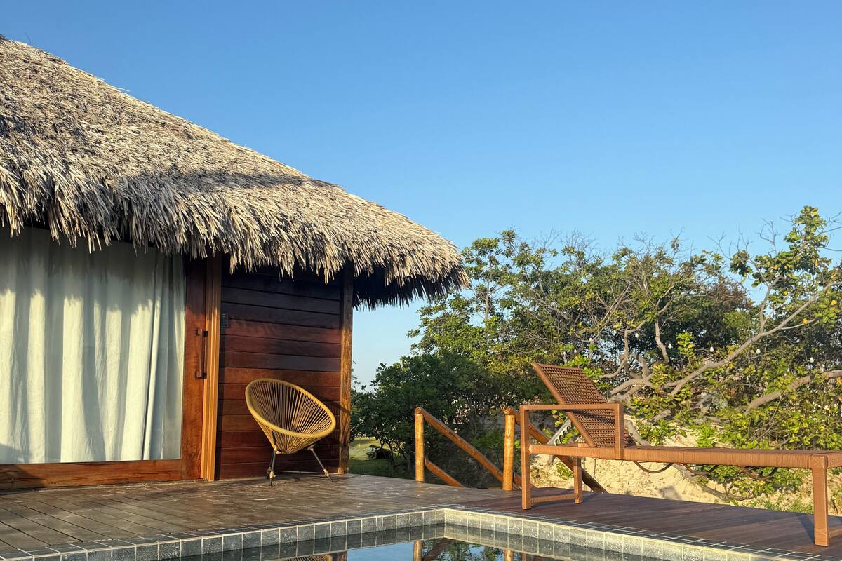 Un área de piscina con terraza de madera, una estructura de techo de paja y una silla de mimbre.