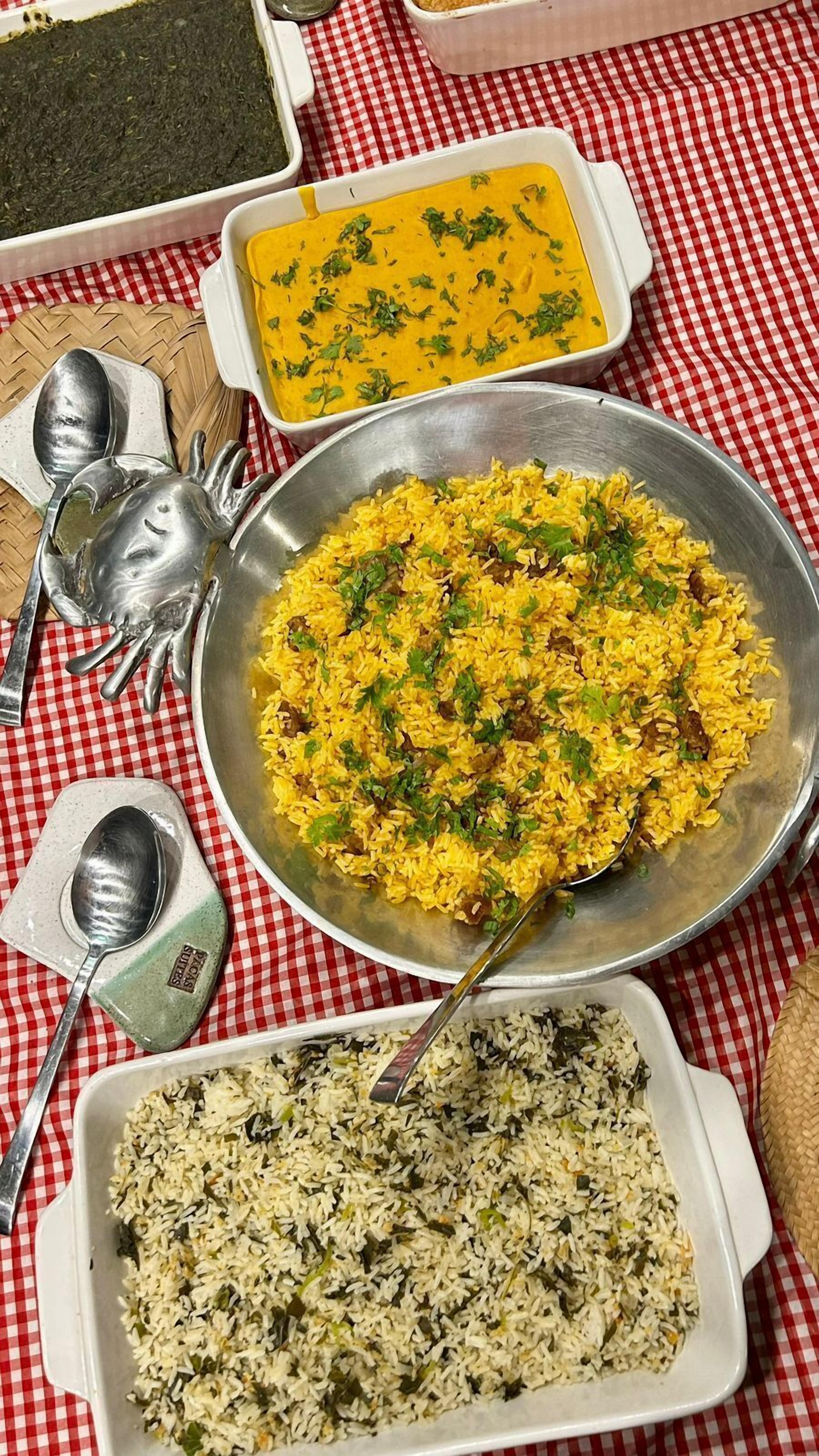 A table with various dishes including yellow rice, a green vegetable dish, and a dark green dish, all on a red and white checkered tablecloth.