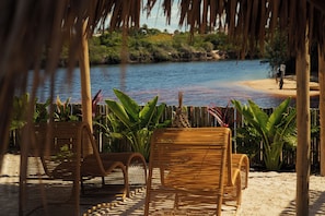 A beachside lounge with wicker chairs and tropical plants, overlooking a calm body of water and a sandy beach. - Pacas Suítes (Santo Amaro do Maranhão)