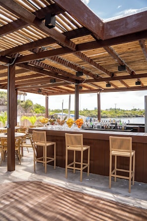 An outdoor bar area with wooden beams, wicker chairs, and a view of the water. - Pacas Suítes (Santo Amaro do Maranhão)