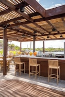 Zona de bar al aire libre con vigas de madera, sillas de mimbre y vista al agua.