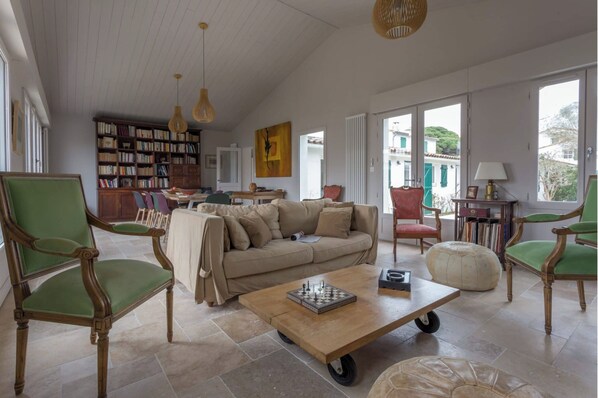 A spacious living room with a large sofa, green armchairs, a coffee table with a chessboard, a bookshelf, and large windows offering a view of buildings. - Villa Luxueuse 4 Chambres Proche Plage / Luxurious Villa 4BR Near Beach (La Couarde-sur-Mer)