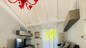 A spacious living room with a wooden floor, a grey sofa, a TV on a low shelf, a red chandelier, and a large window with natural light.