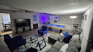Een woonkamer met een pooltafel, blauwe fauteuils, een bank, een televisie en een plafondventilator.
