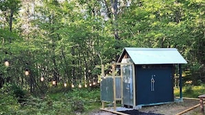 A rustic wooden cabin with a metal door and a blue roof, surrounded by dense greenery and a path leading to it. - Bourbon St BlueBird Bus + HOT TUB on 10 ACRES (BItely)