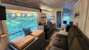 A compact, modern interior with a sofa, a bench with cushions, a small kitchen area, and a view of greenery through large windows. - Bourbon St BlueBird Bus + HOT TUB on 10 ACRES (BItely)