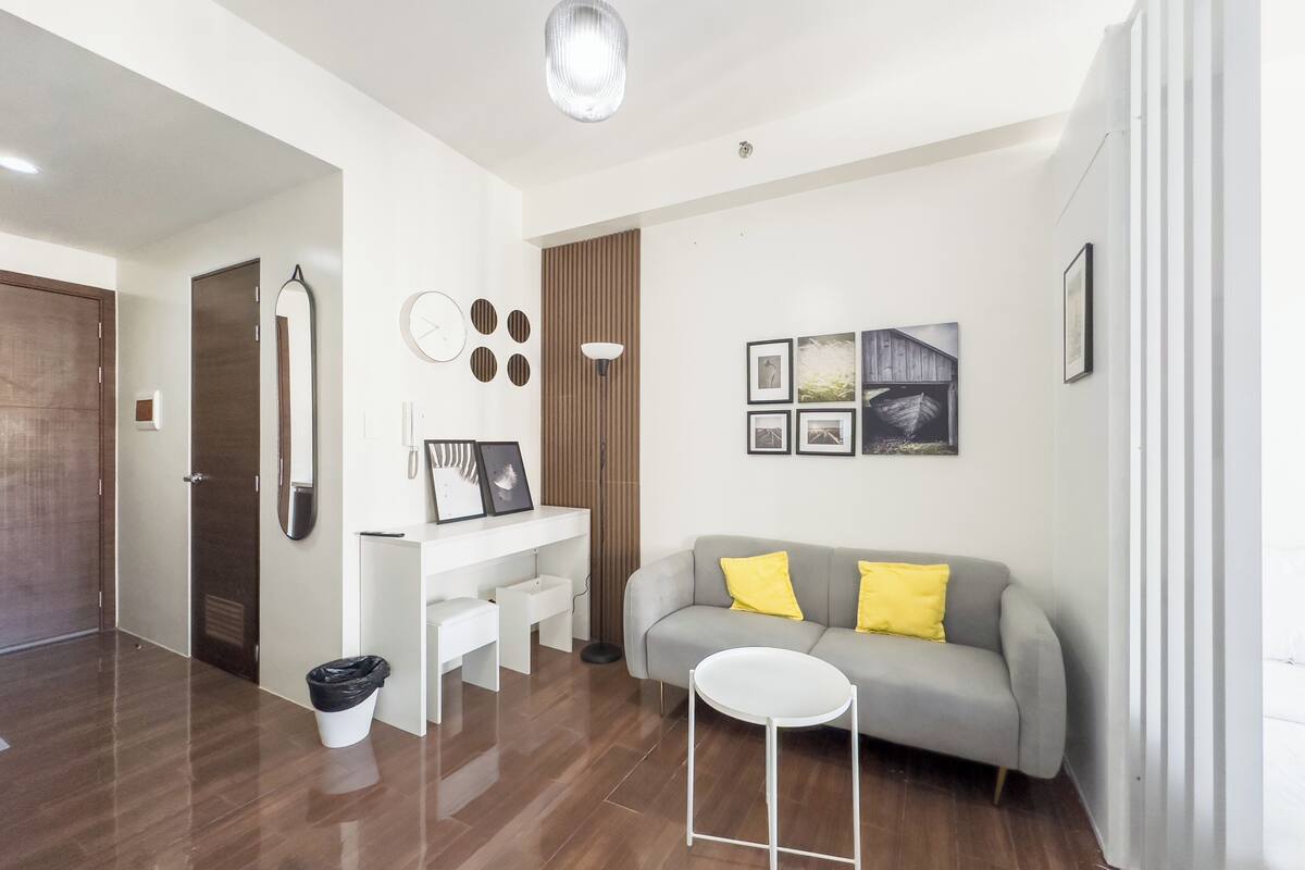 A modern living room with a grey sofa, a white coffee table, a white desk with a mirror, and a large window.