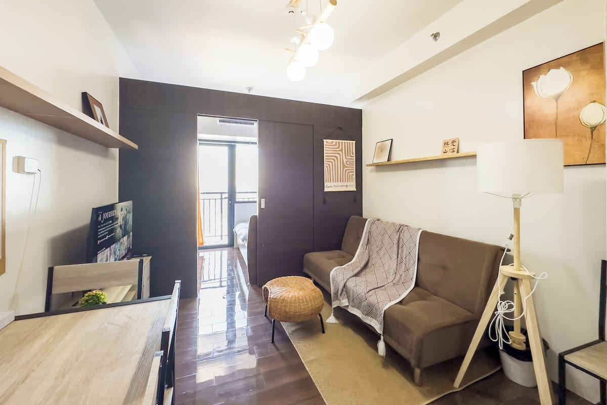 A modern living room with a sofa, a dining table, and a lamp. There is a bookshelf and a painting on the wall.