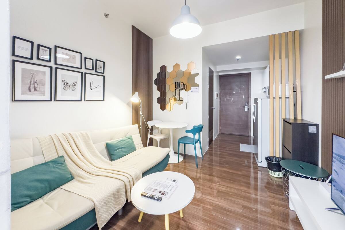 A modern living room with a sofa, a coffee table, and a TV. There are framed pictures on the wall, a dining area, and a kitchen in the background.