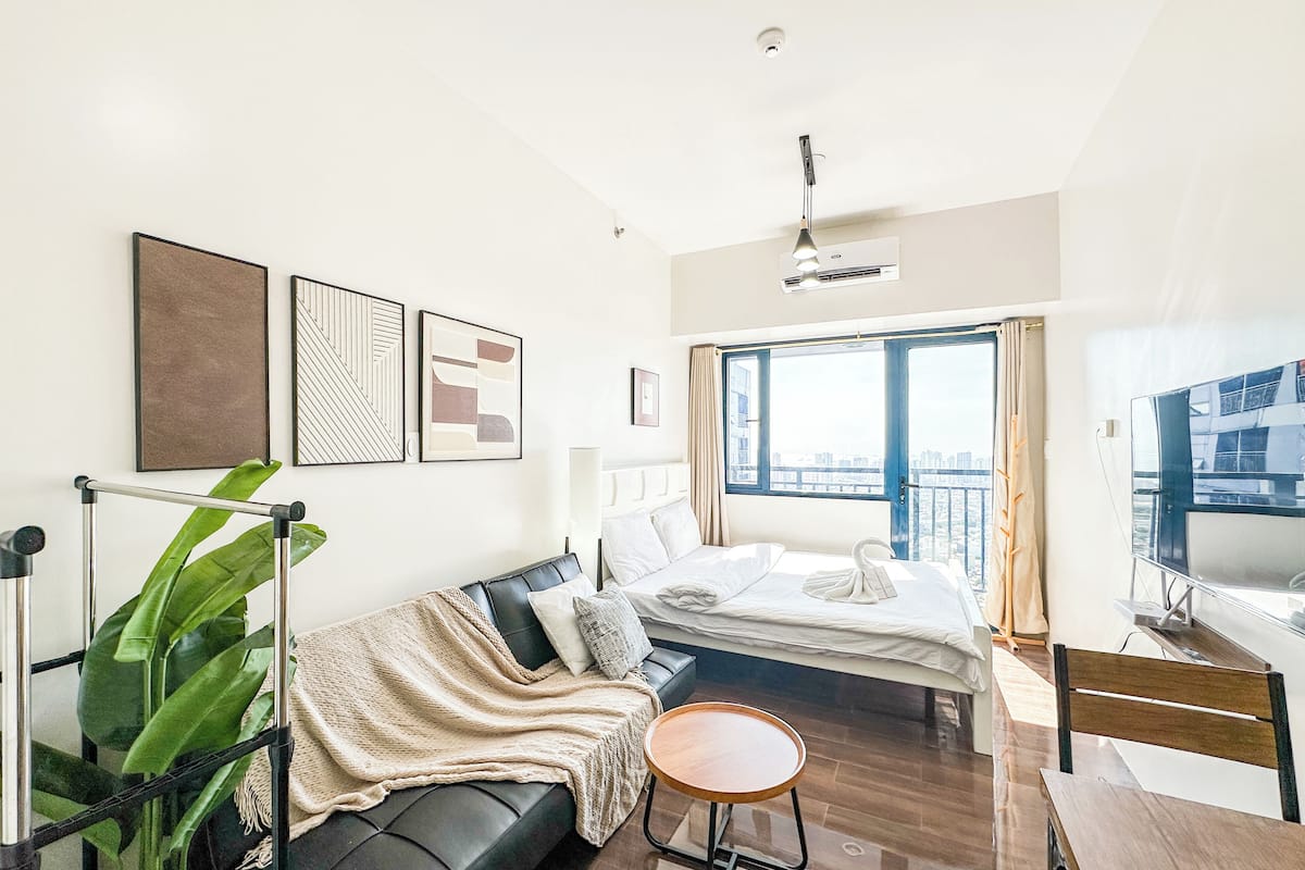 A modern hotel room with a bed, a sofa, a small round table, and a large window offering a city view.