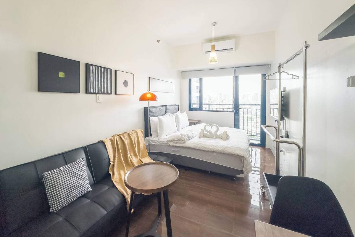 A modern hotel room with a bed, a sofa, a small table, and a balcony with a city view.