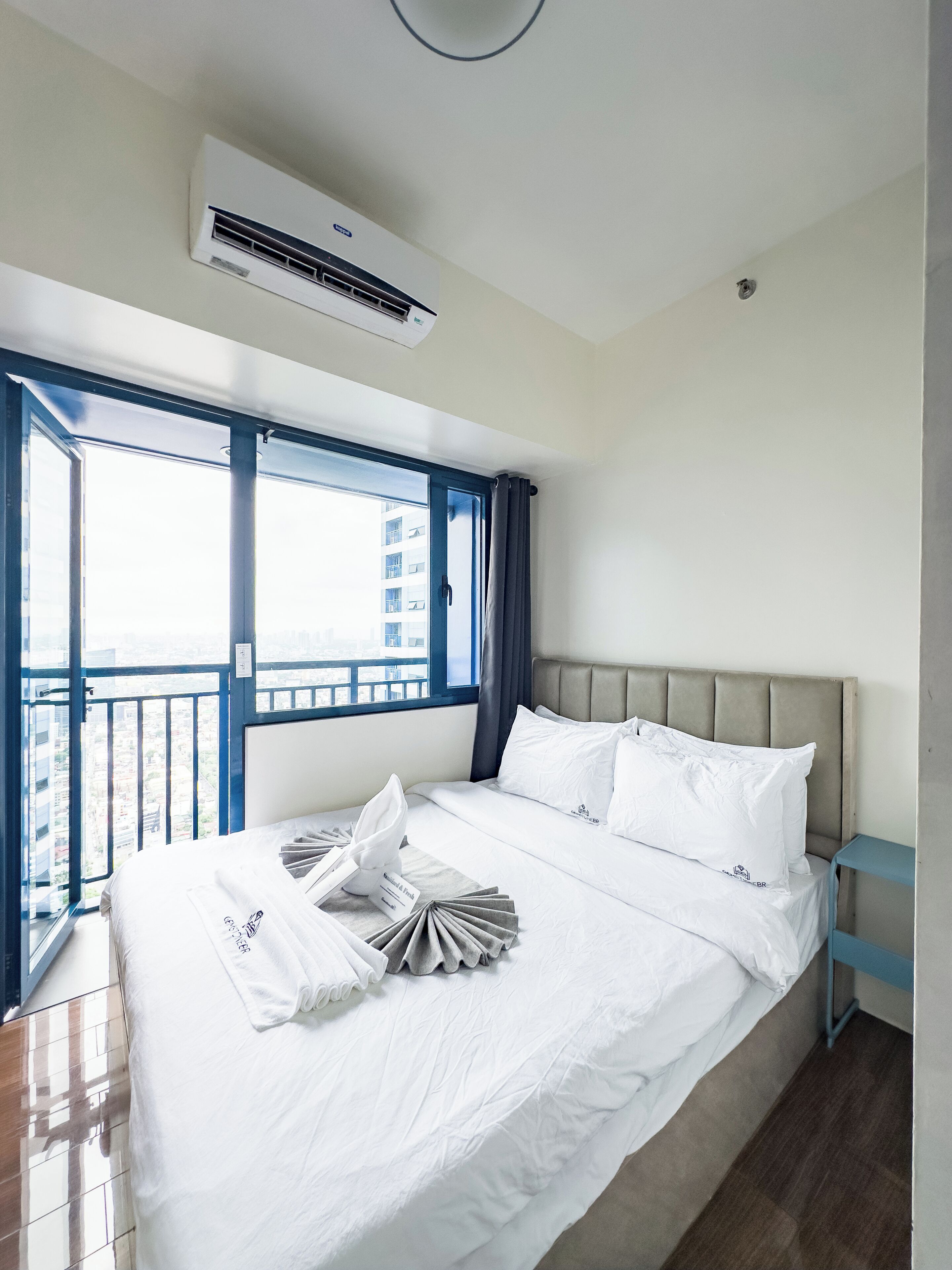A hotel room with a bed, a window with a view of buildings, and an air conditioning unit.