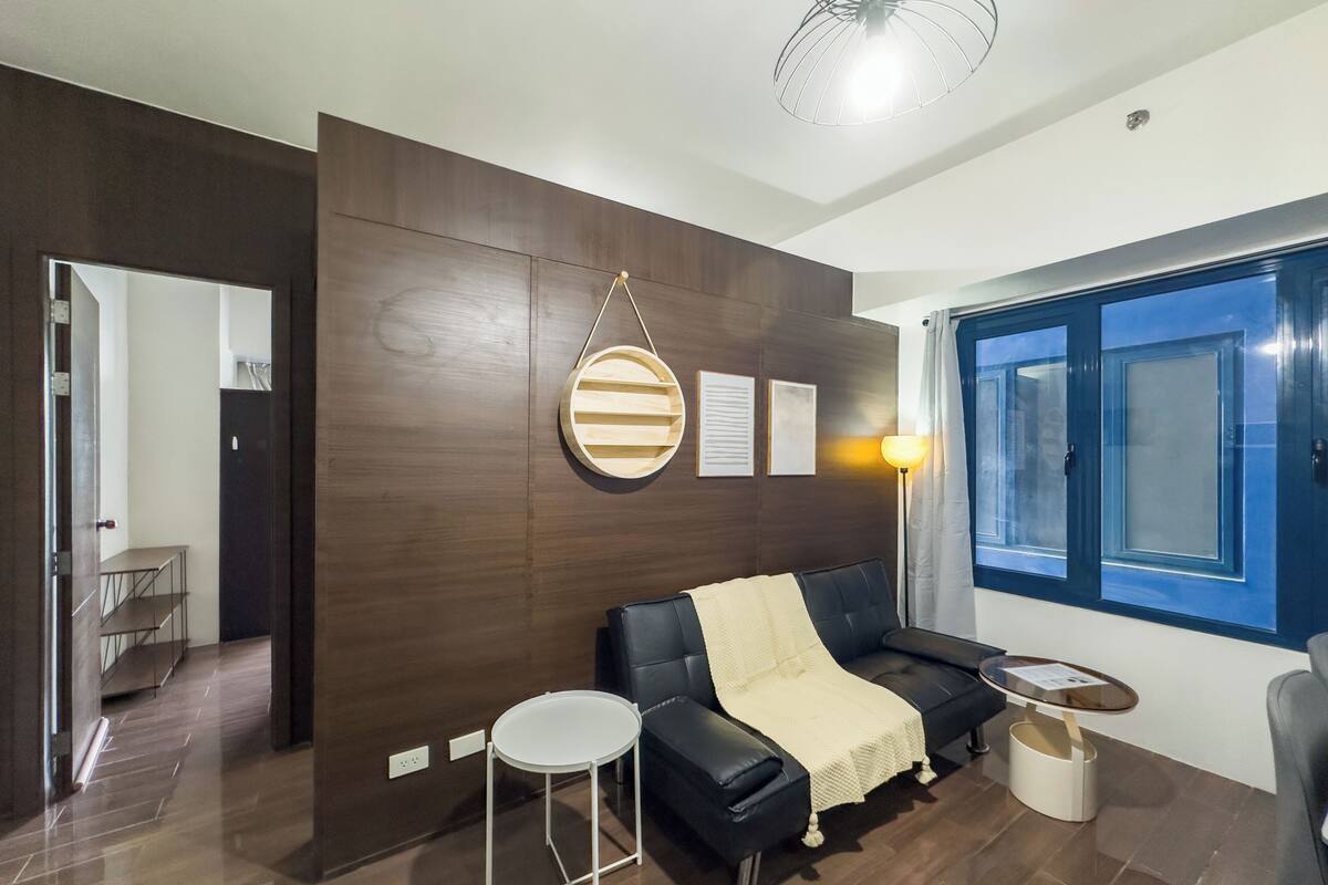 A modern living room with a dark wood accent wall, a black leather sofa, a round wooden shelf, a small white table, and a floor lamp.