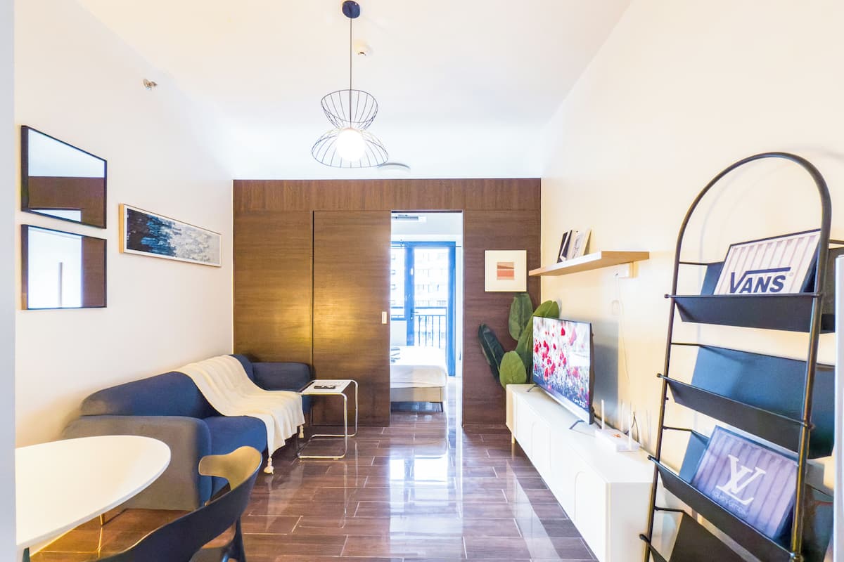 A modern living room with a sofa, a dining table, and a TV. There is a wooden wardrobe and a plant.