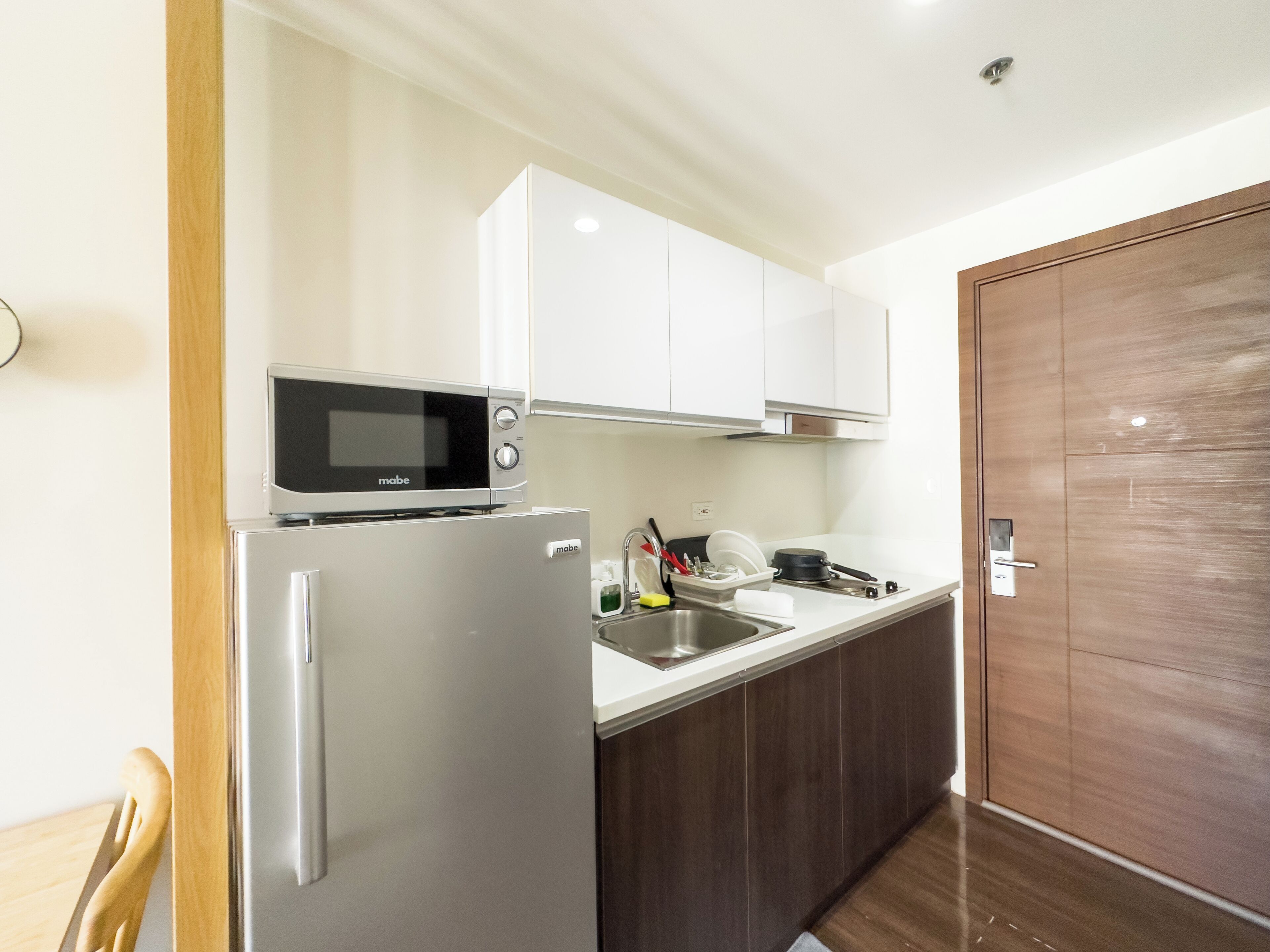 Cocina pequeña con microondas, refrigerador, lavamanos y armarios de madera.