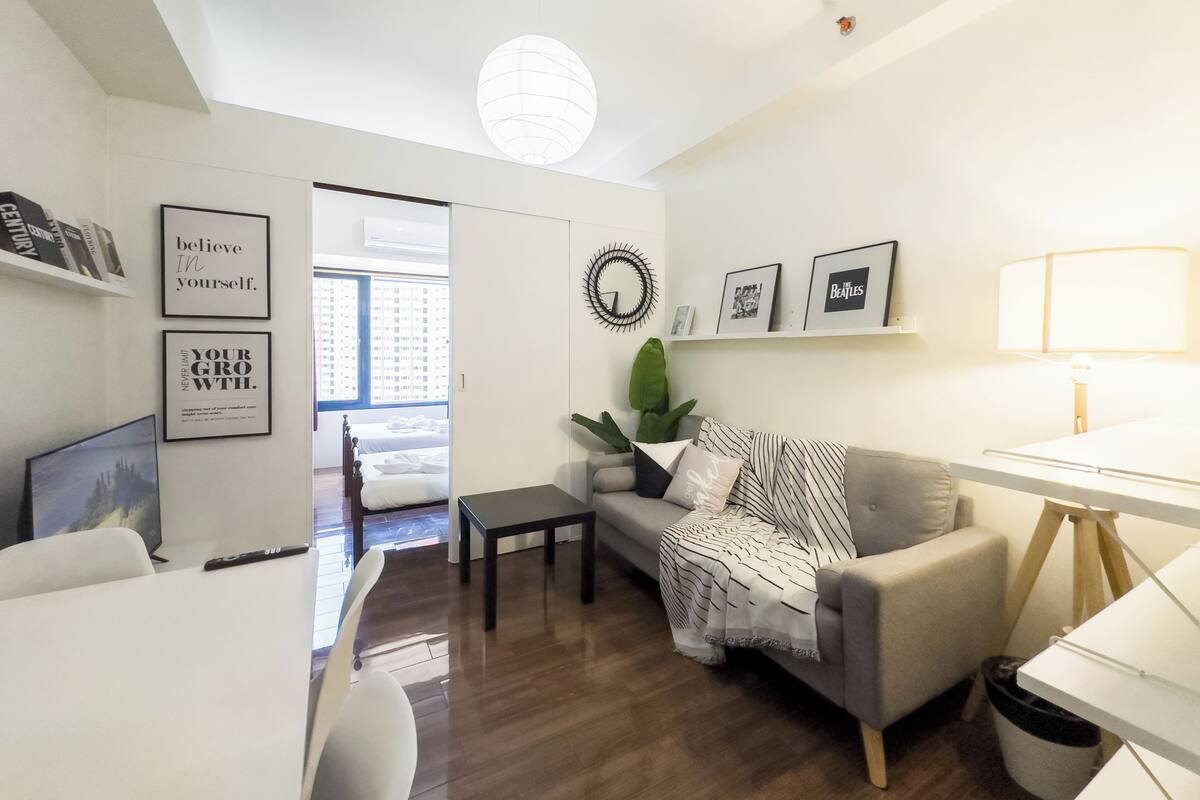 A modern living room with a grey sofa, a small table, and a TV. There are framed pictures on the wall and a plant on the sofa.