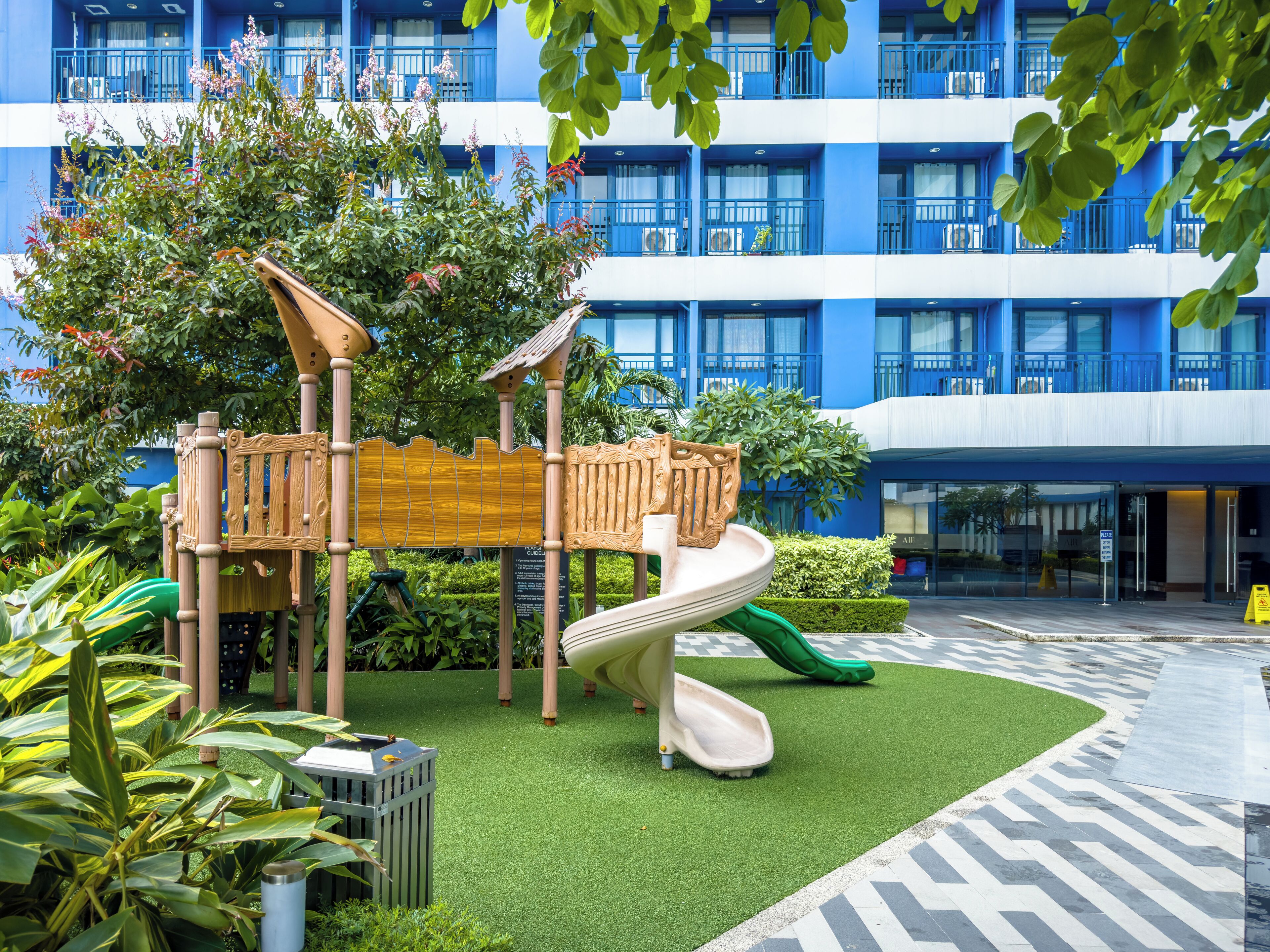 A children's play area with a slide and climbing structures, surrounded by greenery and a blue building in the background.