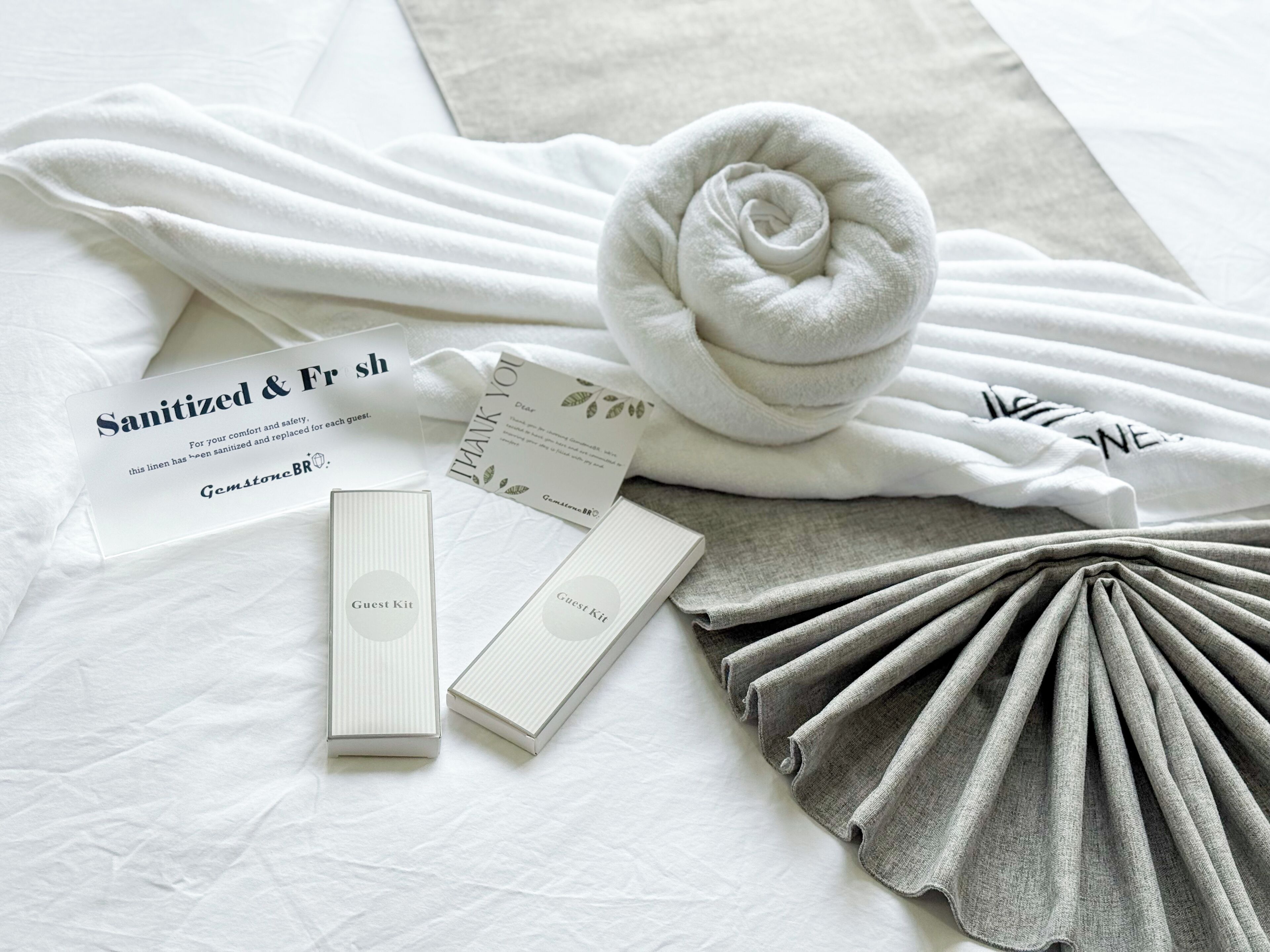 Habitación de hotel ordenada con una toalla blanca doblada, una toalla enrollada, una caja de amenidades para huéspedes y una tarjeta de agradecimiento sobre la cama.