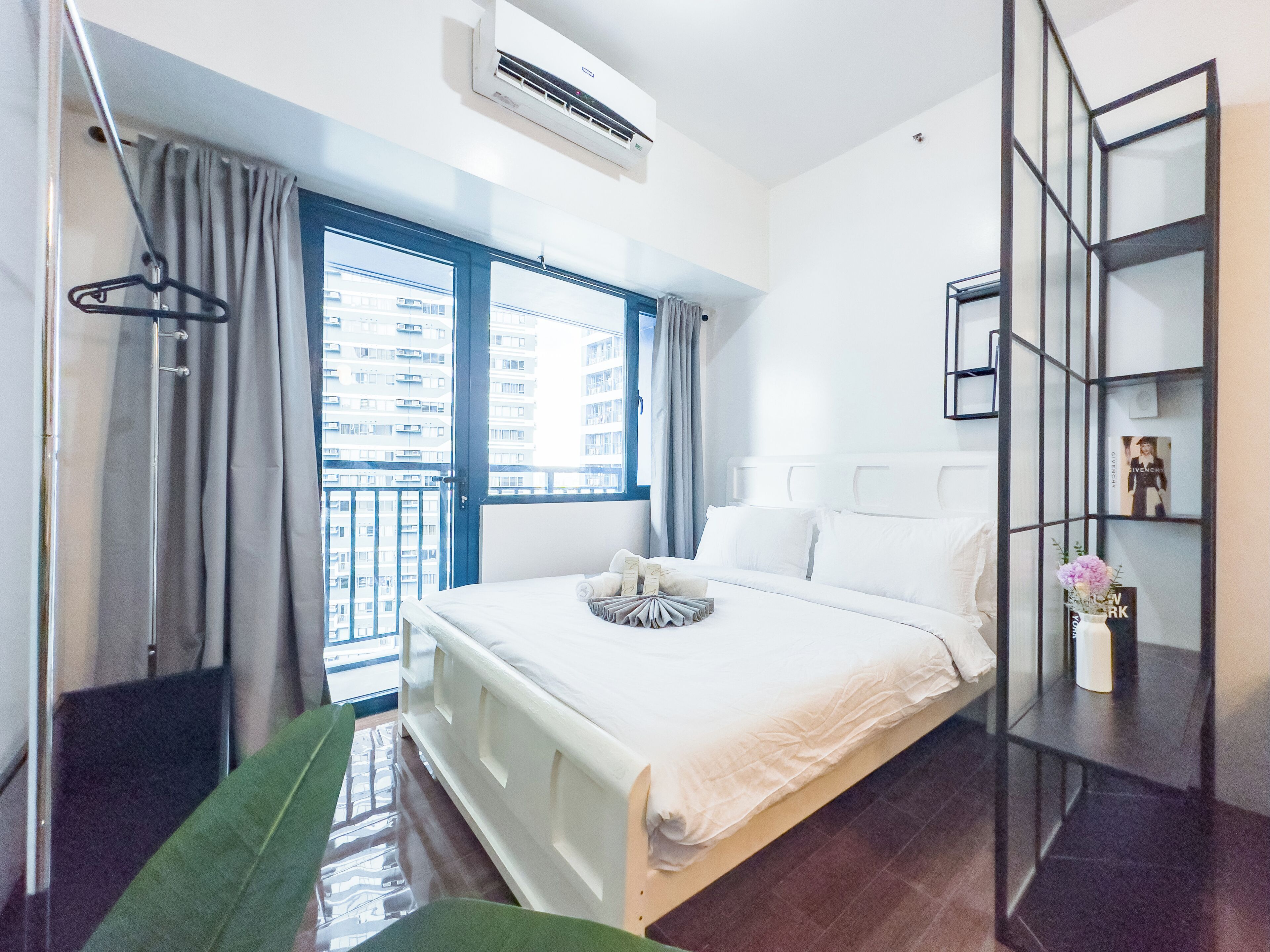 Une chambre d’hôtel avec un lit, une fenêtre avec des rideaux, un climatiseur fixé au mur et une petite étagère sur laquelle se trouve un vase.