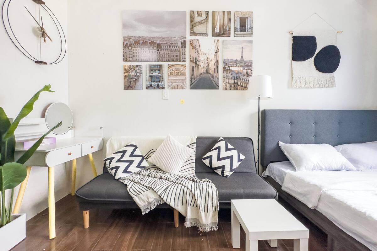 A modern bedroom with a grey sofa, a white bedside table, and a bed with white bedding. There are four framed pictures on the wall, a round mirror on the wall, and a plant in a white pot.