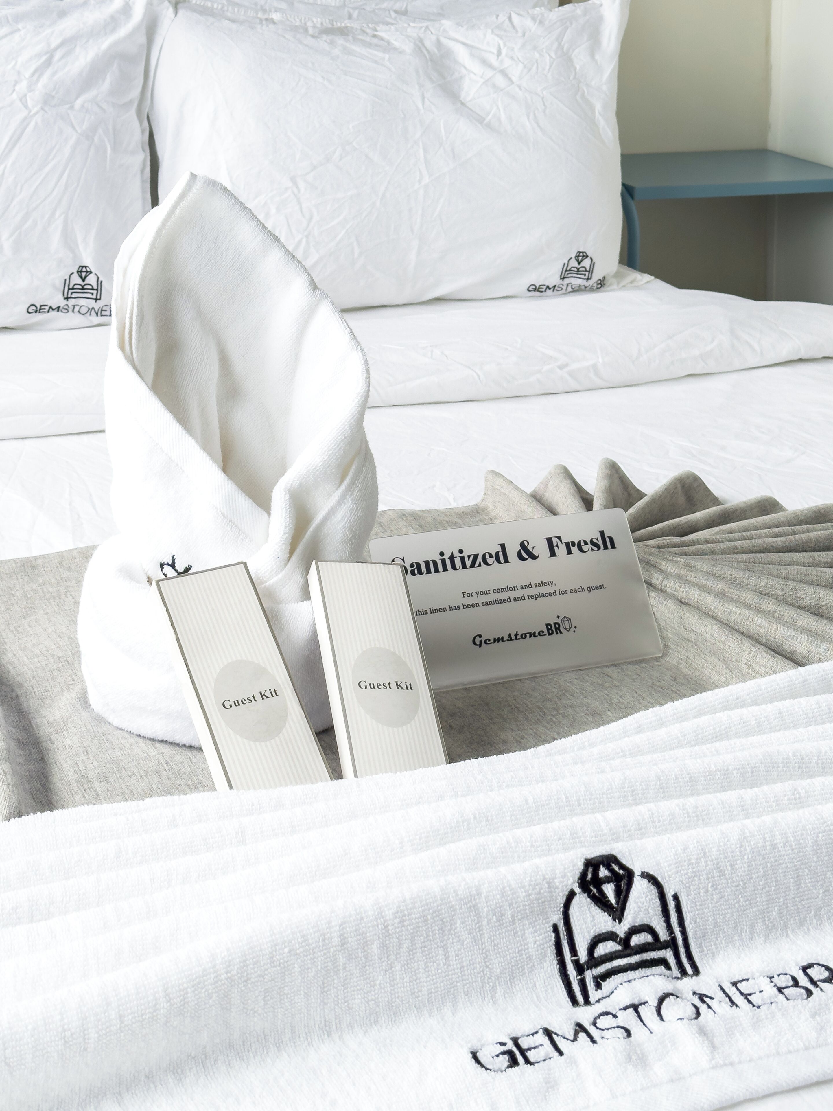 A neatly arranged bed with a white pillow, a folded towel, and a guest kit including a sanitizer and a tissue.