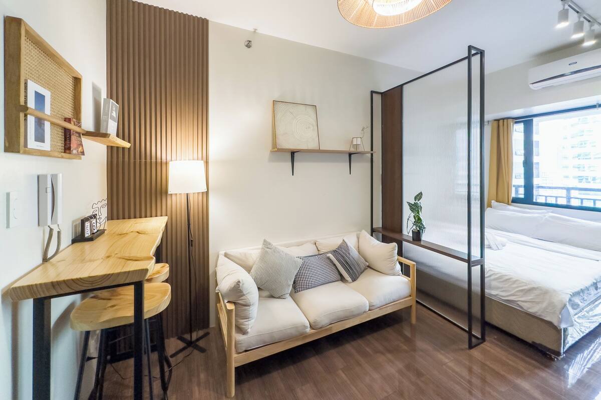 A modern hotel room with a wooden desk, a sofa, a bed, a large window with a view, and a ceiling light fixture.