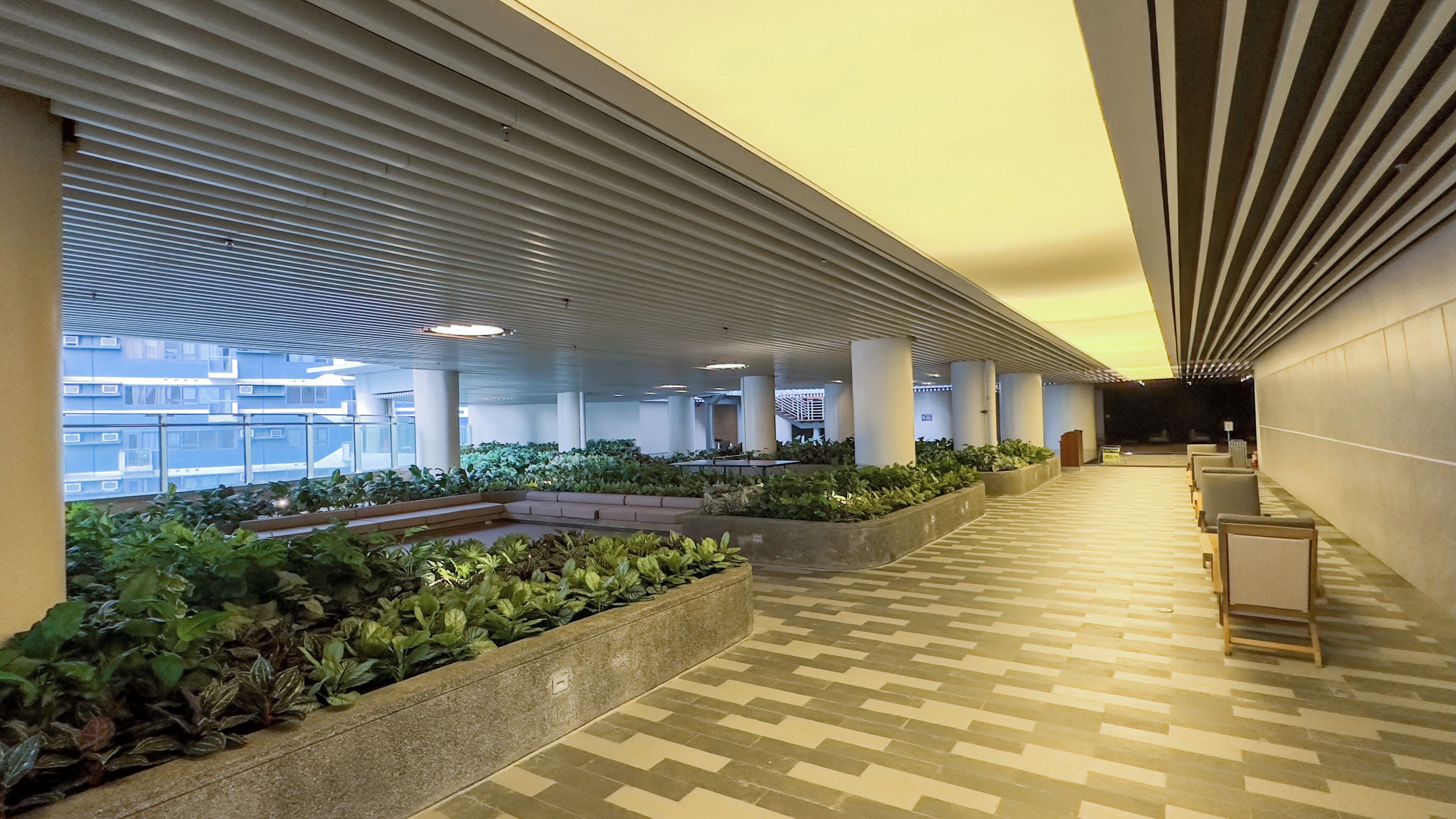 A corridor with a patterned floor, planters with greenery, and a seating area with chairs.