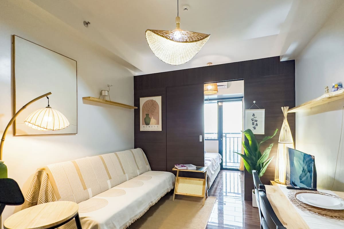 A modern living room with a sofa, a dining table, and a TV. There are decorative items on shelves and a large window providing natural light.