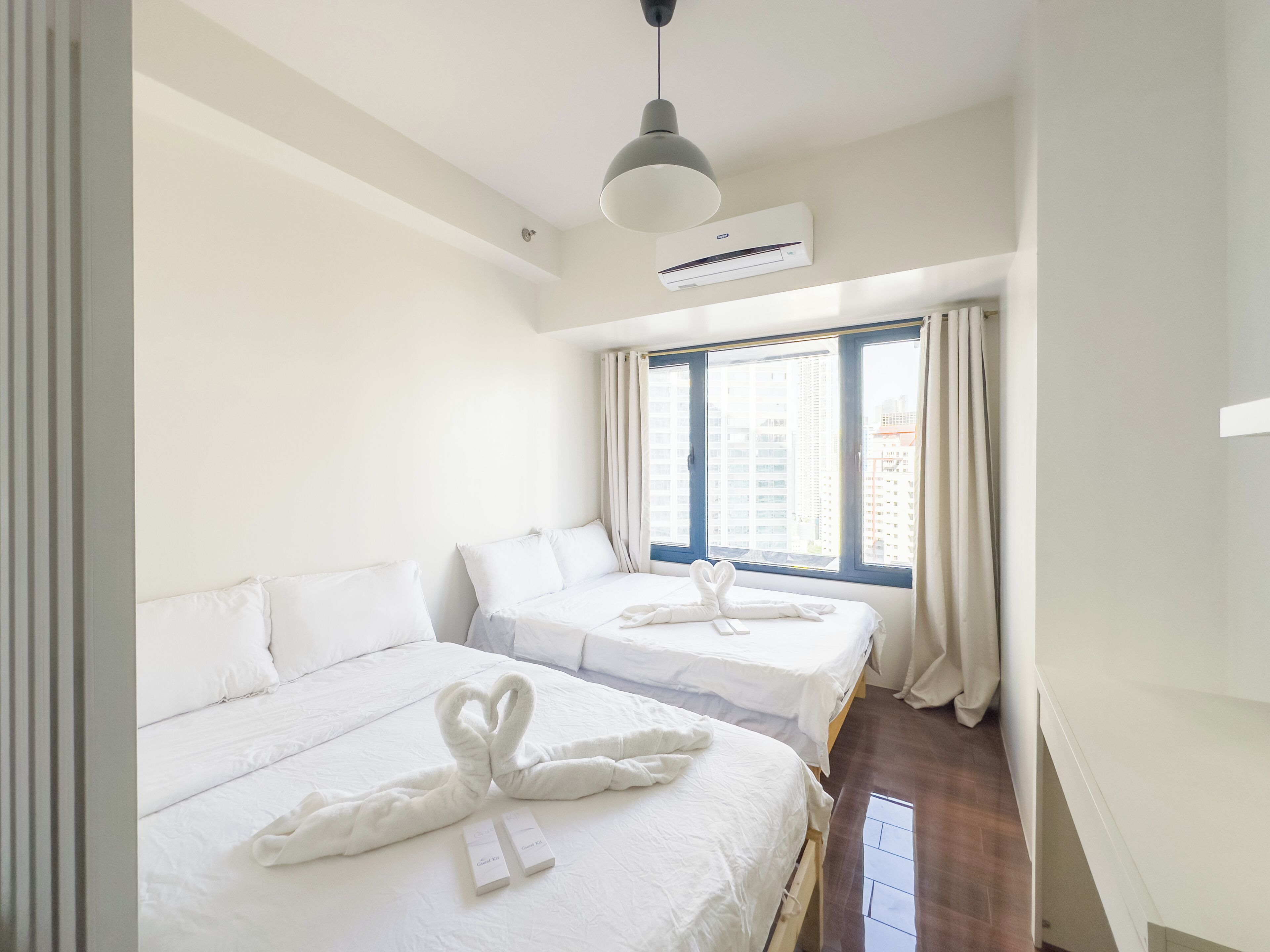 A hotel room with two beds, a window with curtains, an air conditioning unit, and a pendant light.