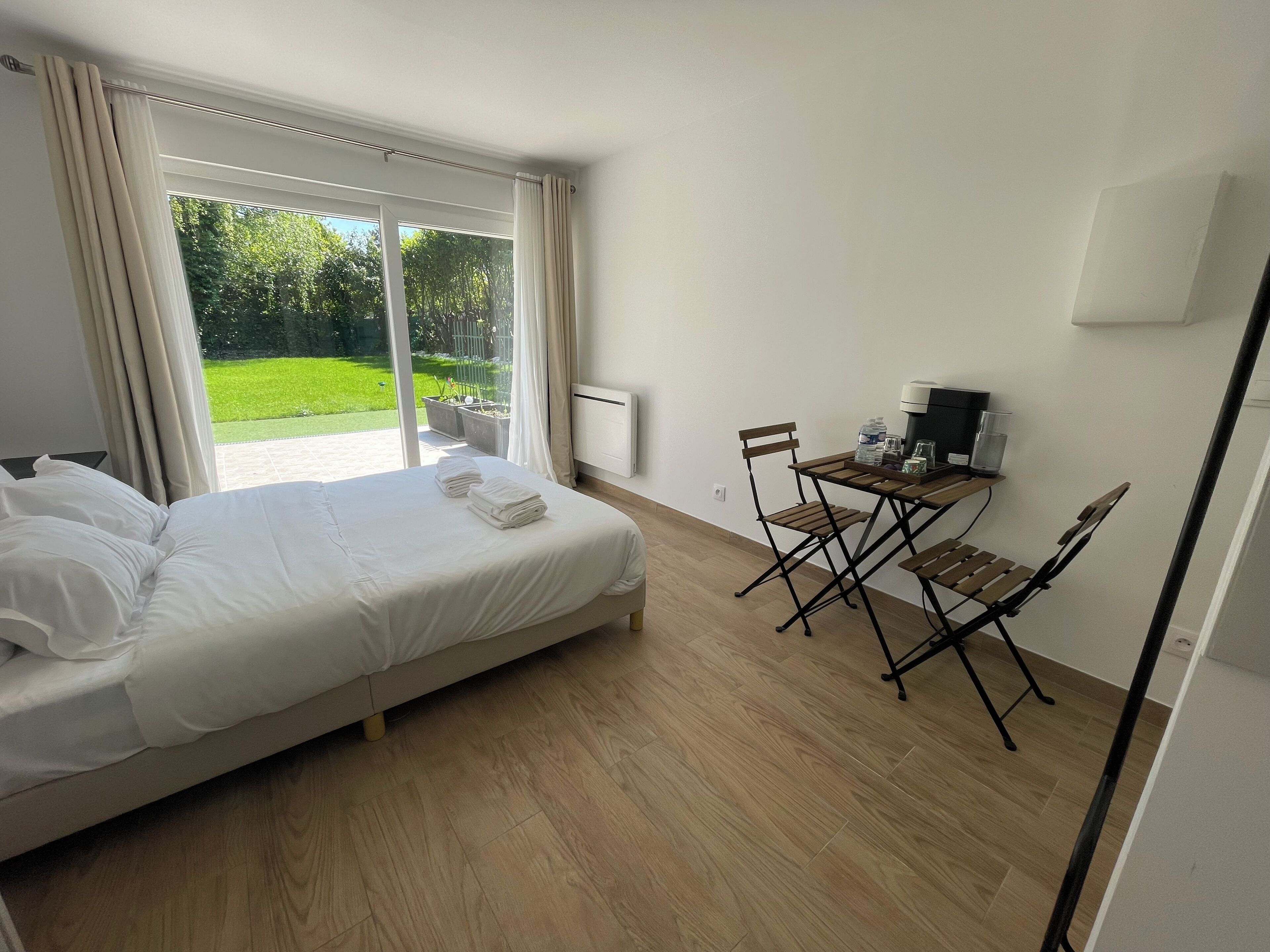 Une chambre avec un lit, une petite table et des chaises, une machine à café et une vue sur un jardin verdoyant, visible à travers une grande fenêtre.