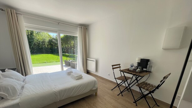 Un dormitorio con cama, una pequeña mesa con sillas, una cafetera y vistas a un jardín verde a través de un ventanal grande.