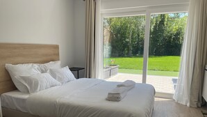 Una cama bien hecha con sábanas blancas y una toalla doblada, una mesita de noche pequeña y vistas a un jardín verde a través de un ventanal corredero.