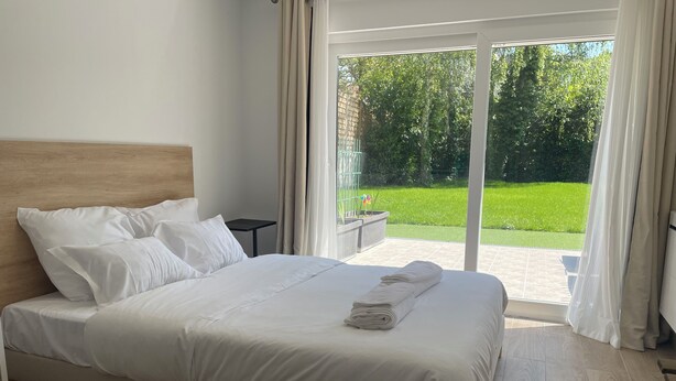 A neatly made bed with white linens and a folded towel, a small bedside table, and a view of a green garden through a sliding glass door. - Koalawakeup (Vitry-sur-Seine)